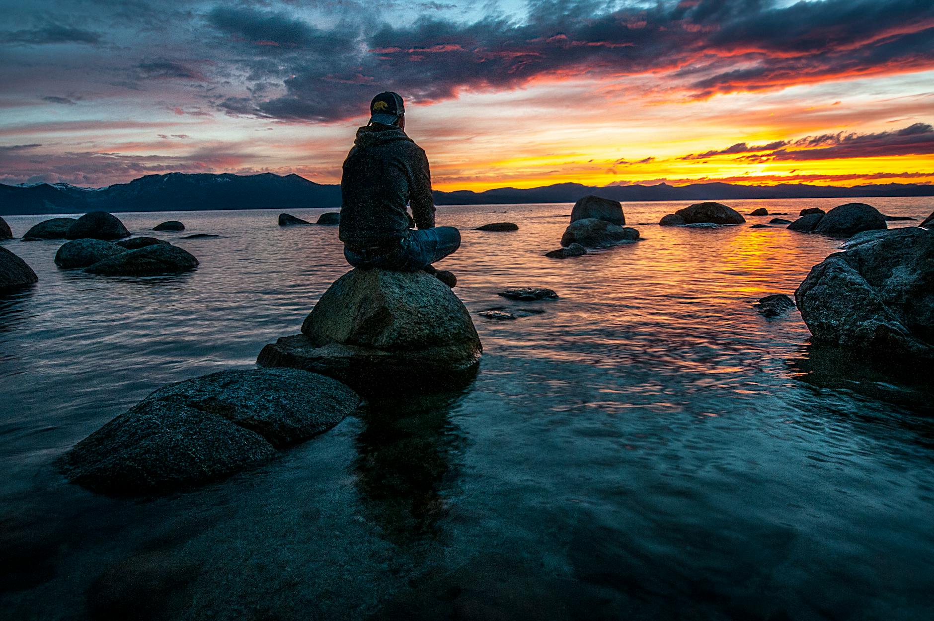 A man sitting on a rock by the ocean as the sun sets. He is thinking.