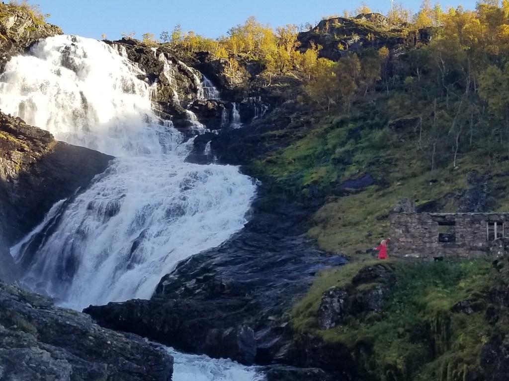 A waterfall in the mountains. A woman in a red dress is standing to the right of the waterfall | Scandinavian Folklore