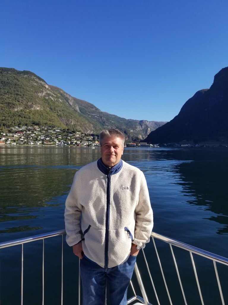 The photo is of me standing on the deck of a boat in Sognefjord. There are mountains and a village in the background. I am wearing a white jacket.