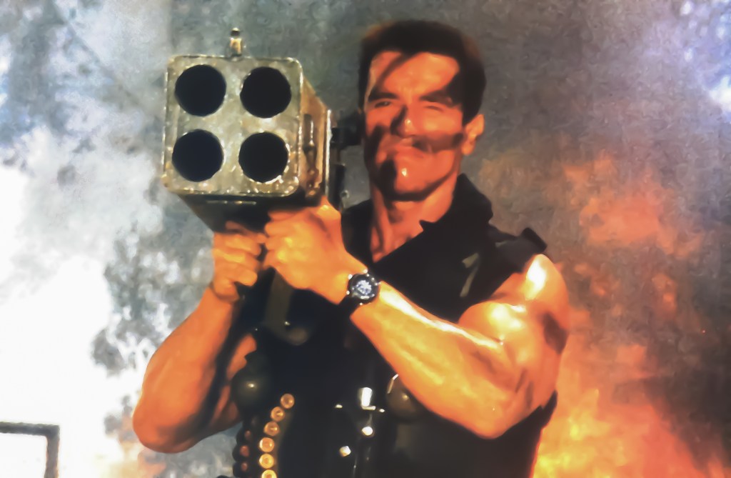 Arnold Schwarzenegger is holding a gian machine gun pointed into the camera. There is fire and smoke in the background.