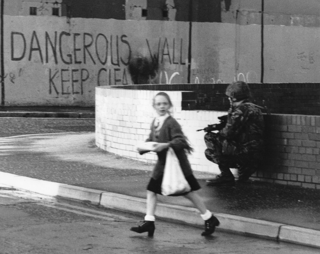 Black and white photo showing a young girl crossing the street as an armed soldier is hunkered down behind her. There is big wall with text written on it saying, “Dangerous Wall keep Clear.”