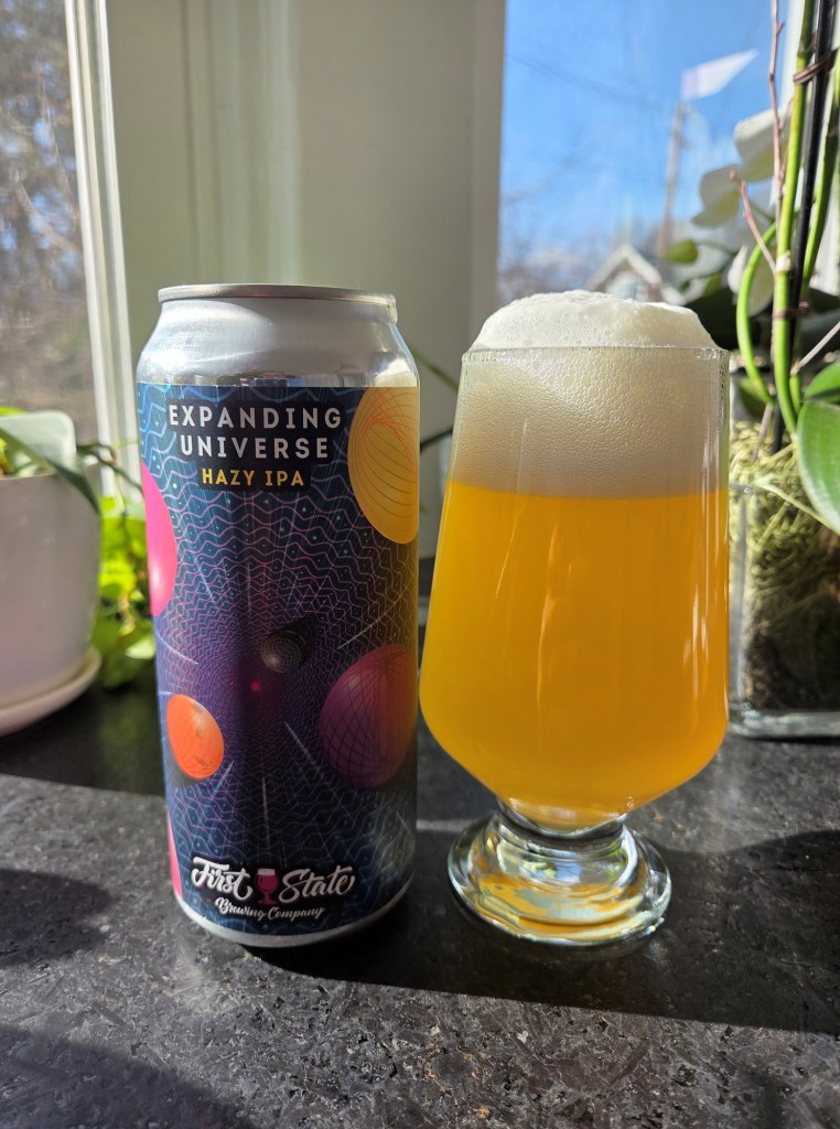 The picture shows a glass with yellow to light orange hazy liquid. There are green plants in the background.