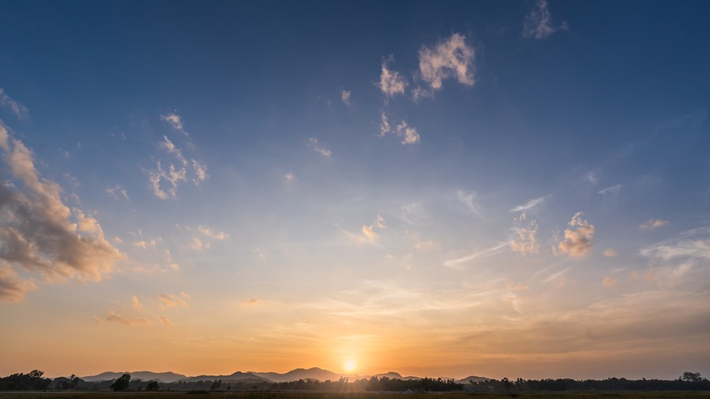 The sun is setting over distant mountains. There are some clouds, a forest, and field in the foreground. The sun has a yellow tint; the clouds are slightly yellow-orange and sky above is blue.